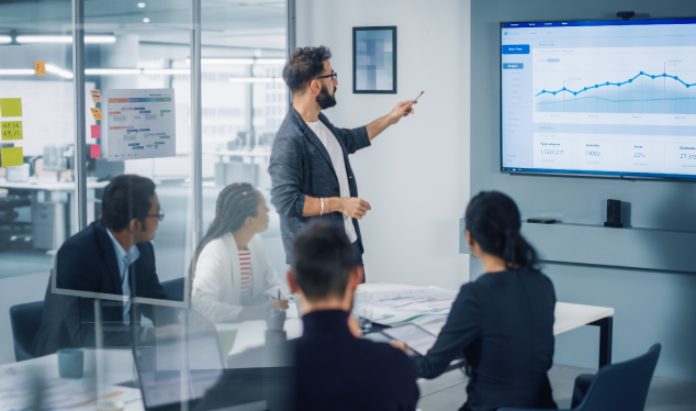 Corporate meeting in a modern conference room with glass walls. A bearded man wearing glasses points to a large screen displaying a growth chart, while four colleagues seated at the table pay close attention. The environment is professional and collaborative.