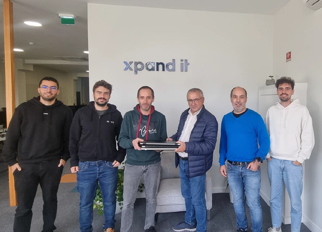 Grupo de seis pessoas posa para a foto numa sala da empresa Xpand IT, com dois homens ao centro a segurar uma pilha de laptops. A imagem simboliza doação de equipamentos tecnológicos e responsabilidade social corporativa.