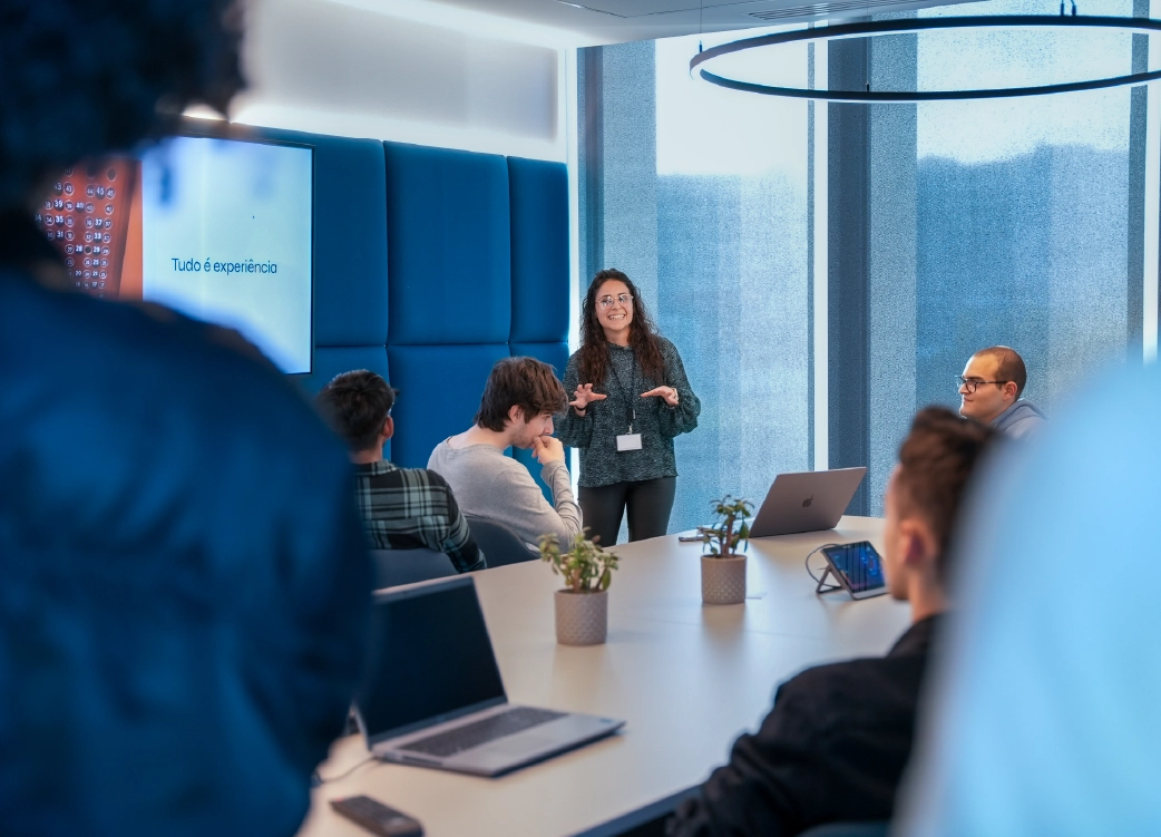 A woman leads a training session in a modern corporate setting, with attentive participants gathered around a table equipped with laptops and work materials. The image represents innovation, continuous learning, and professional collaboration.