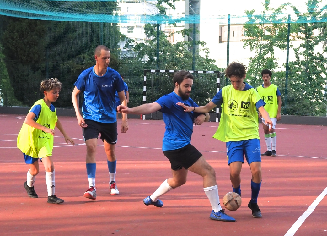Jovens e adultos jogam futebol num campo ao ar livre, com coletes de diferentes cores para distinguir as equipas. A imagem representa desporto, inclusão social e espírito de equipa.