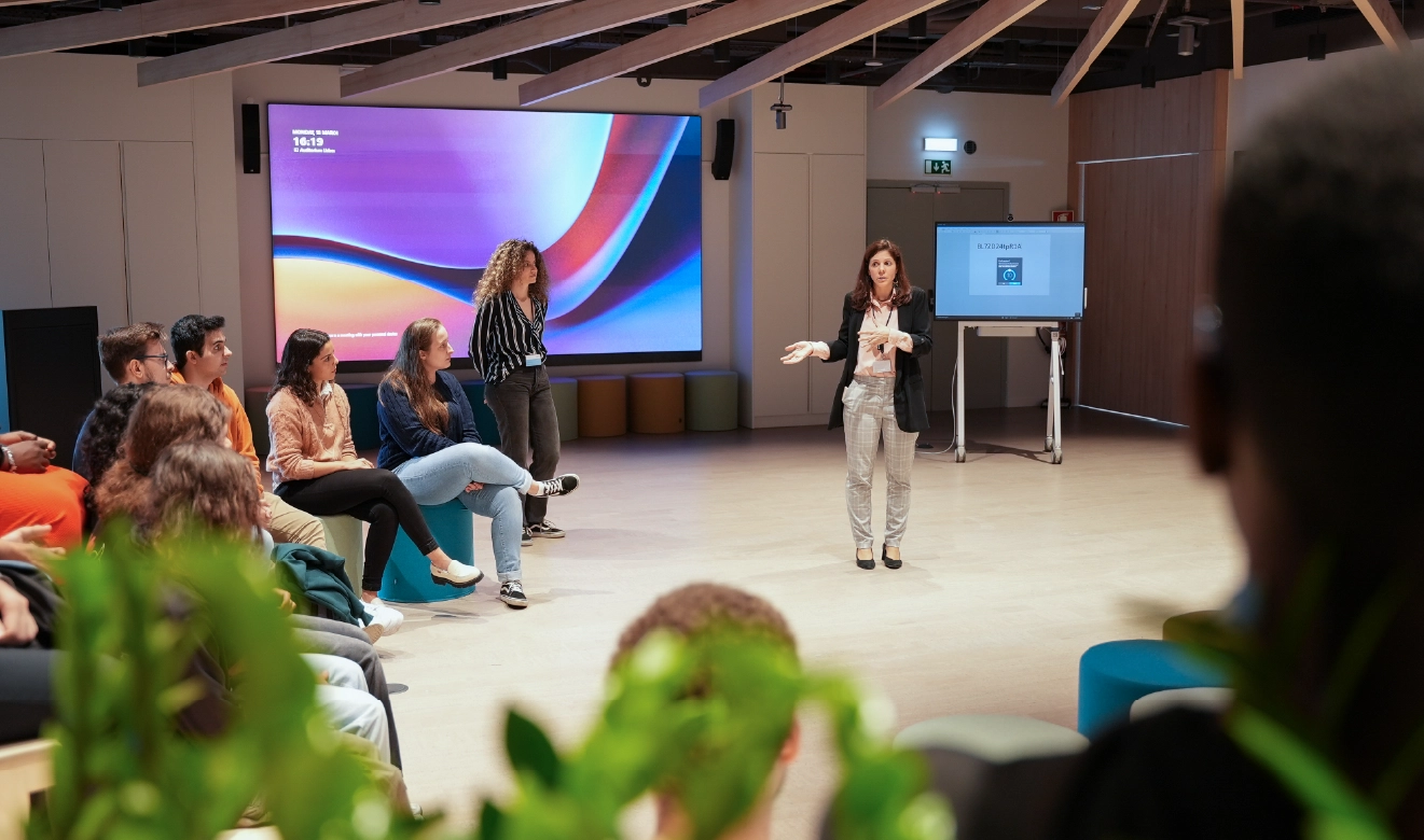 Uma mulher conduz uma apresentação interativa para um grupo de jovens atentos, num espaço moderno com um ecrã digital de fundo. O ambiente sugere educação, inovação e responsabilidade social.