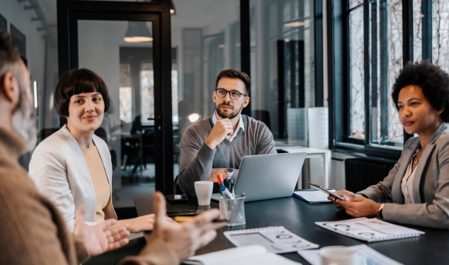 A group of four professionals gathered in a modern conference room, discussing business strategies. A man wearing glasses listens attentively while others interact, conveying an atmosphere of collaboration and decision-making.