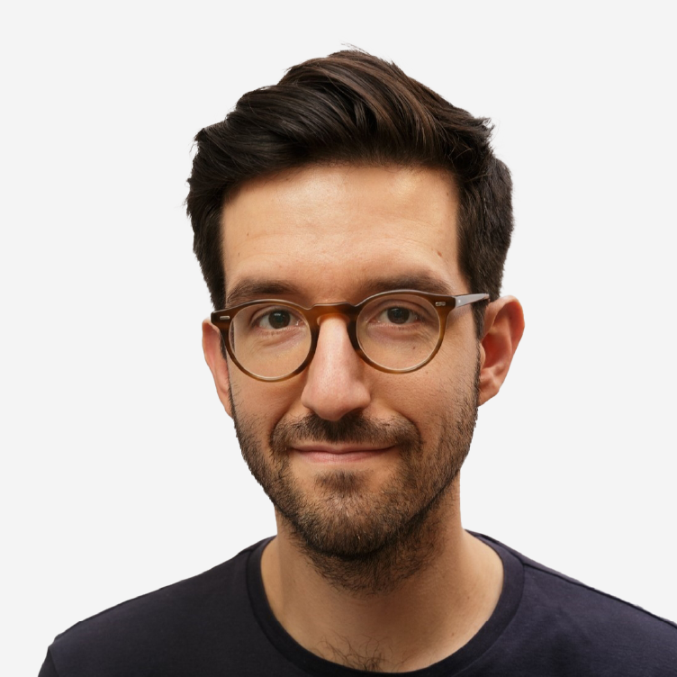 Retrato de um homem com óculos de massa, cabelo castanho escuro, barba curta e t-shirt escura, sobre fundo branco.