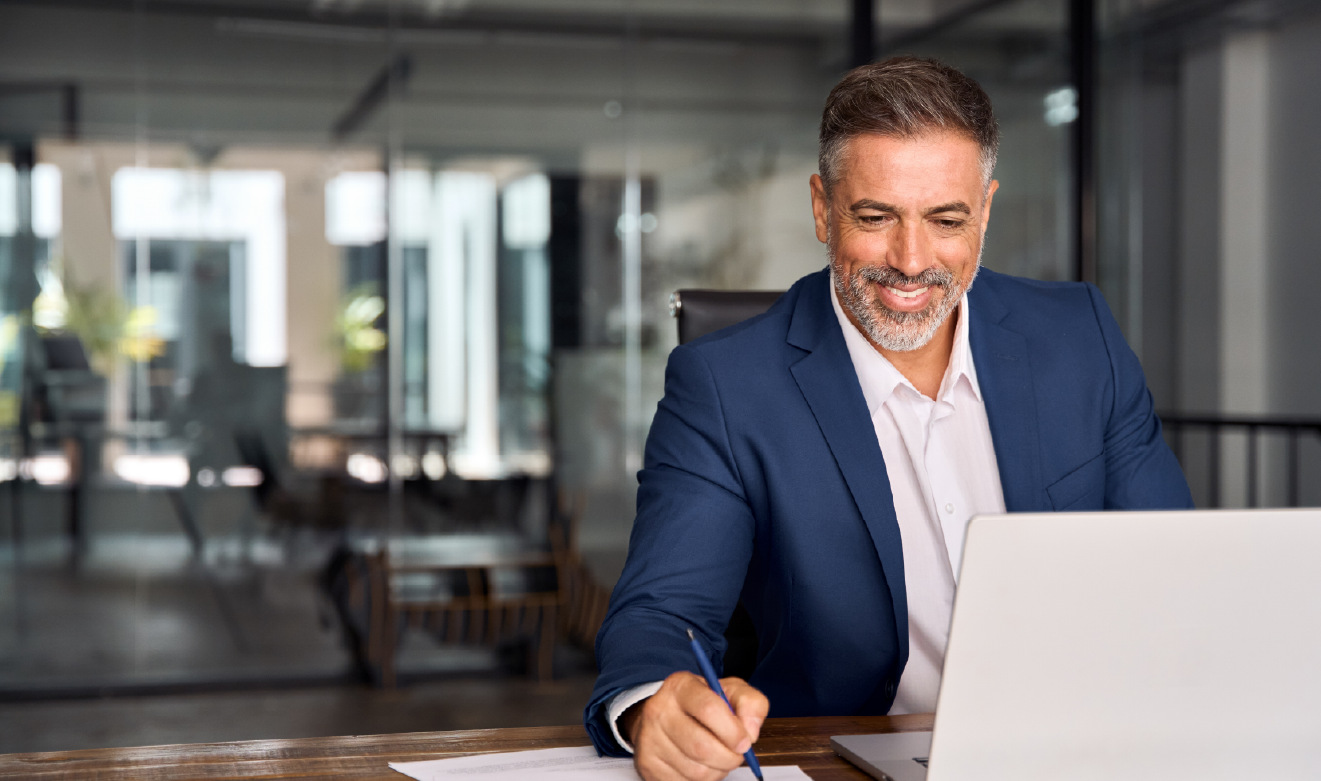 Homem de meia-idade, bem vestido com fato azul e camisa branca, a sorrir enquanto trabalha num computador portátil num ambiente de escritório moderno, sugerindo envolvimento profissional em tecnologias digitais.
