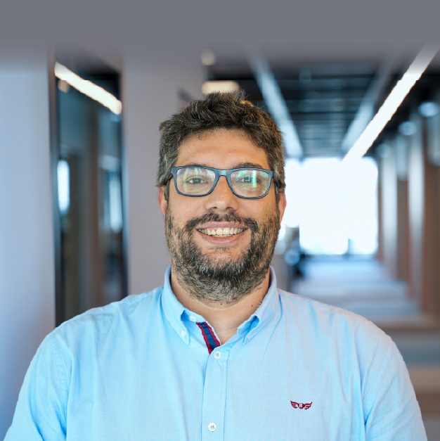 Retrato profissional de um homem sorridente, com óculos, num ambiente empresarial moderno, destacando inovação e tecnologia.