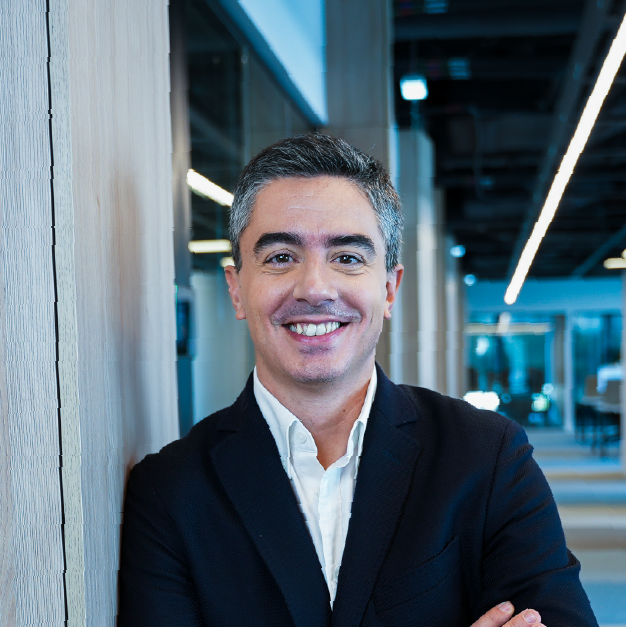 Retrato profissional e sorridente de um homem de fato num contexto empresarial moderno e tecnológico.