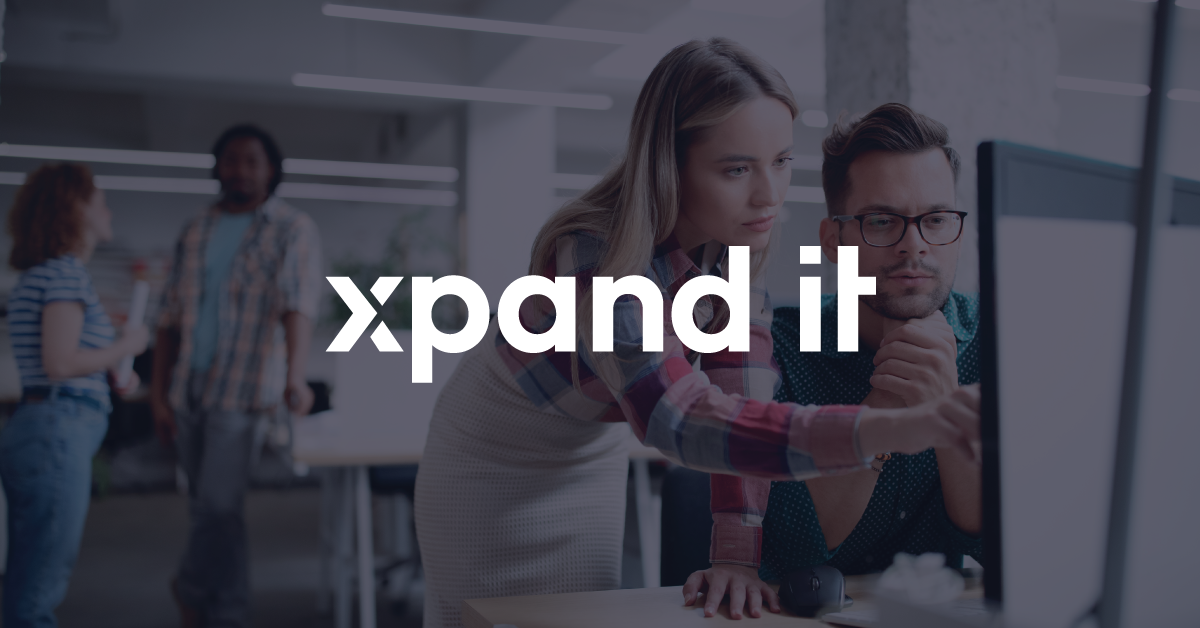 Two colleagues closely examine a monitor while discussing a tech solution in a modern office setting, with the “Xpand IT” logo prominently displayed.