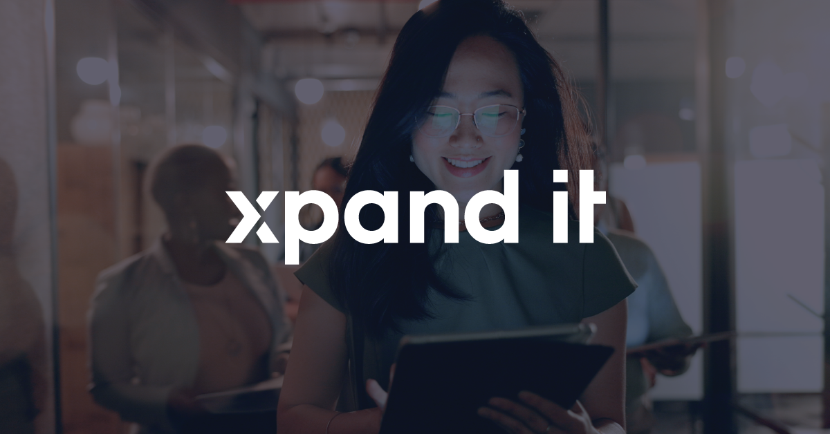 A smiling woman holding a tablet in a well-lit hallway, with the "Xpand IT" logo overlaid at the center. In the background, other people walk through the corporate environment, conveying innovation and digital transformation.