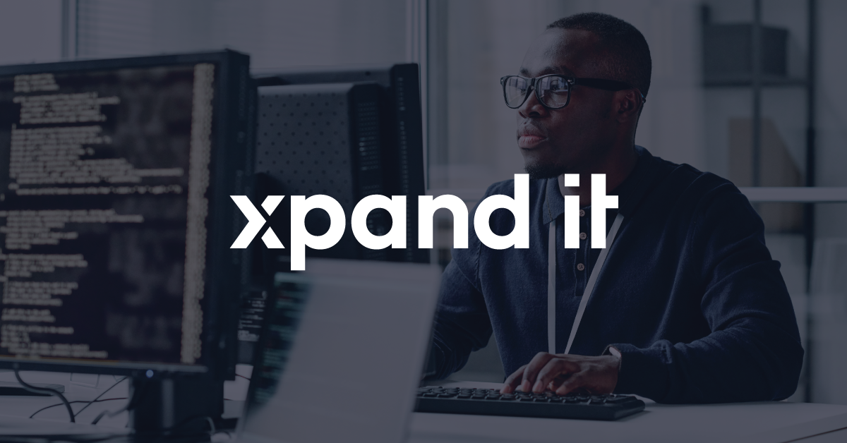 Desenvolvedor full-stack num escritório tecnológico, a trabalhar num computador enquanto olha para um monitor com código, com o logótipo "Xpand IT" sobreposto.