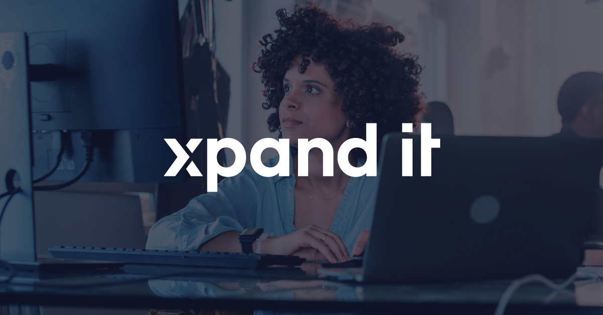 Female developer working in a modern office environment with dual monitors and a laptop, with the Xpand IT logo prominently centered. The image symbolizes focus, innovation, and system integration.