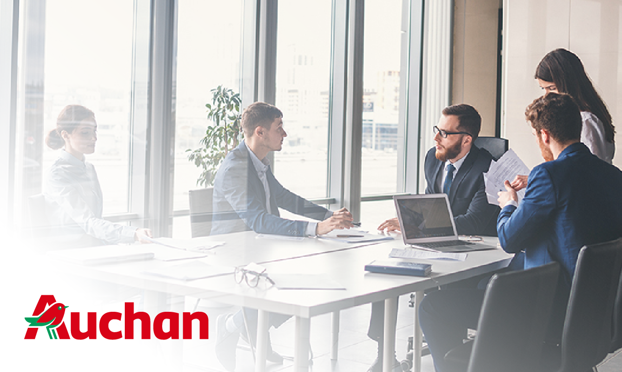 Equipa de profissionais em reunião numa sala de reuniões moderna e luminosa, com o logótipo da Auchan em destaque. A imagem representa colaboração estratégica e sucesso empresarial.