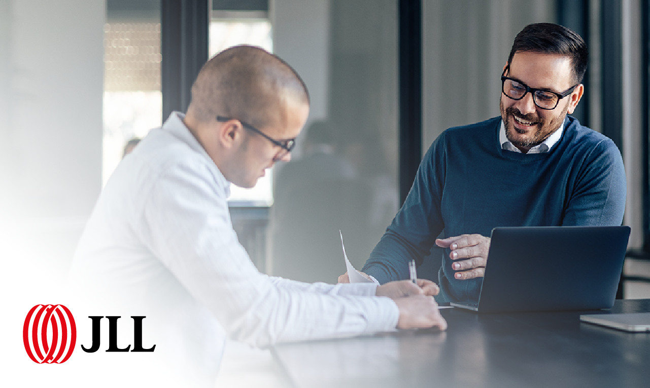 Dois profissionais em conversa num ambiente de escritório moderno, com computador portátil e documentos. O logótipo da JLL transmite confiança, colaboração e sucesso na transformação digital.