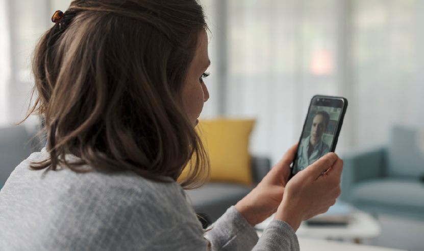 A woman attends a remote medical consultation via video call on her smartphone, with a doctor visible on the screen. The image represents the advancement of telemedicine and digital access to healthcare.