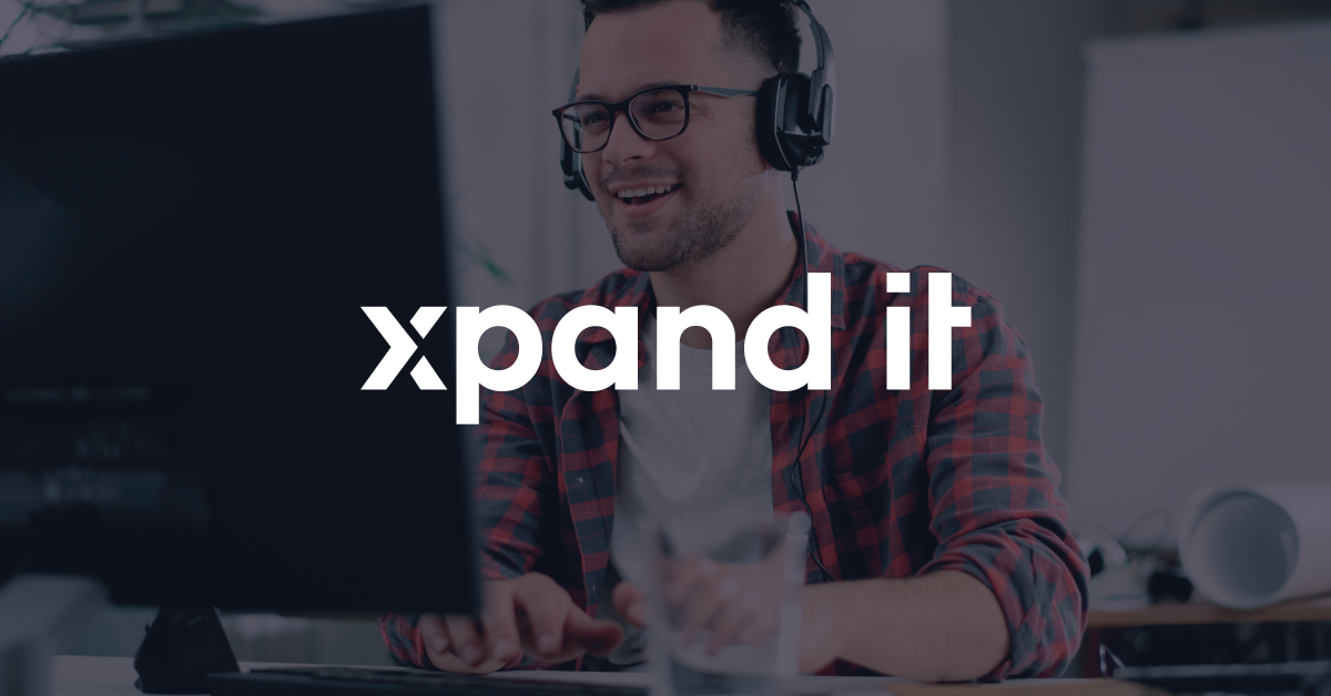 Homem a sorrir com auscultadores e camisa aos quadrados, em frente a um monitor num ambiente de trabalho moderno. O logótipo da Xpand IT está sobreposto no centro da imagem.