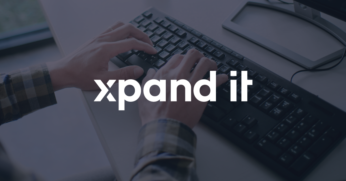 Hands typing on a black computer keyboard, with the Xpand IT logo overlaid in the center. The setting includes a white desk and part of a monitor.