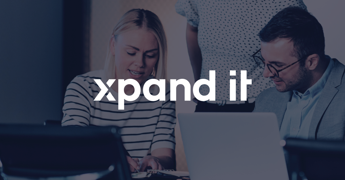 Dois profissionais de tecnologia, uma mulher e um homem, colaboram num ambiente de escritório moderno, focados num portátil, com o logótipo da Xpand IT sobreposto. A imagem transmite inovação, trabalho em equipa e cultura tecnológica.