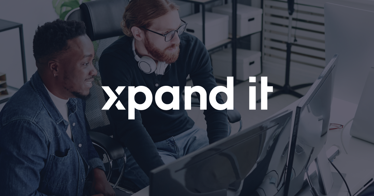 Two developers seated side by side review code on dual monitors, with the Xpand IT logo prominently centered. The image conveys collaboration and focus on digital solutions.