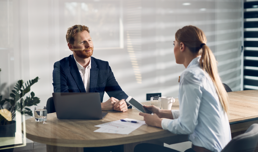 Man and woman in a formal meeting in a modern office, discussing documents on a table with a laptop and tablet, representing negotiation or financial consulting.