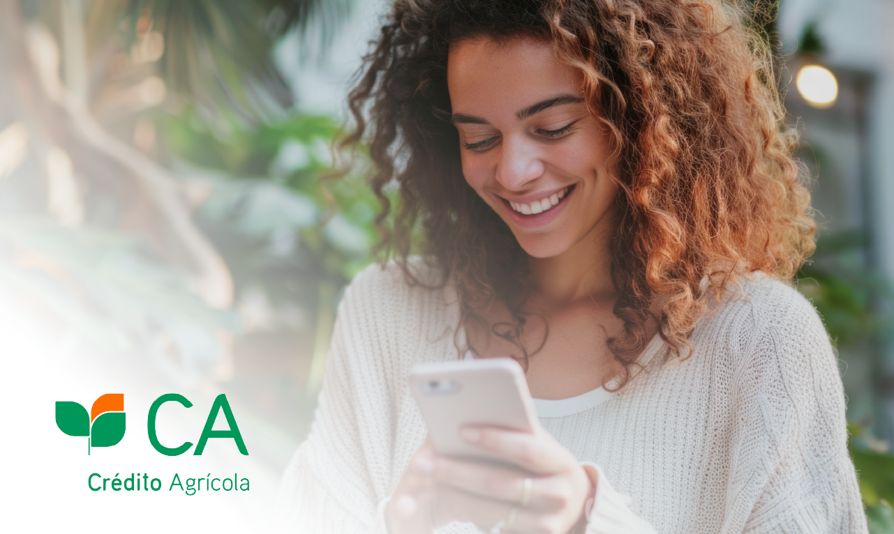 Mulher jovem a sorrir enquanto interage com o seu smartphone, em ambiente iluminado e natural, com o logótipo do Crédito Agrícola no canto inferior esquerdo. Imagem transmite simplicidade, confiança e foco na experiência digital do cliente.