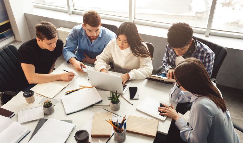 Grupo de jovens estudantes reunidos em torno de uma mesa, colaborando em projetos num portátil, rodeados de cadernos e material de estudo, representando aprendizagem e inovação académica.