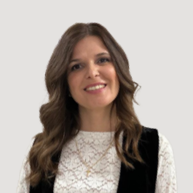 Mulher a sorrir, com cabelo castanho ondulado, vestida com blusa rendada branca e colete preto, em frente a um fundo liso bege