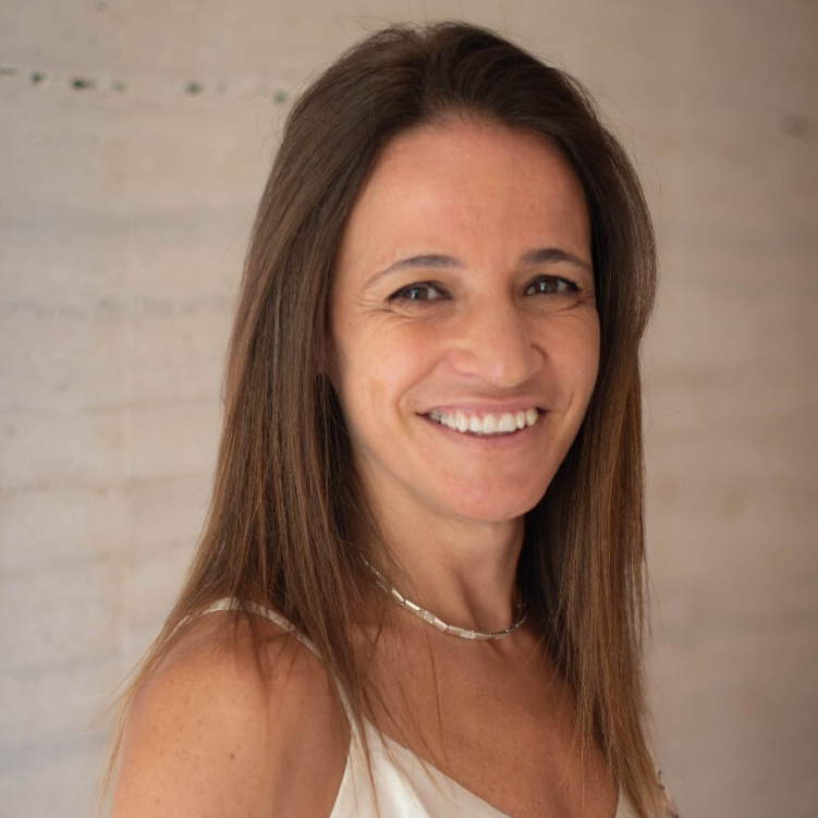 Mulher de cabelo liso castanho, a sorrir com expressão natural, vestida com top claro e colar prateado, em frente a uma parede de betão em tom bege.