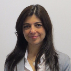 Retrato de uma mulher sorridente com cabelo escuro e liso, vestida com blazer cinzento e camisa branca, sobre fundo neutro.