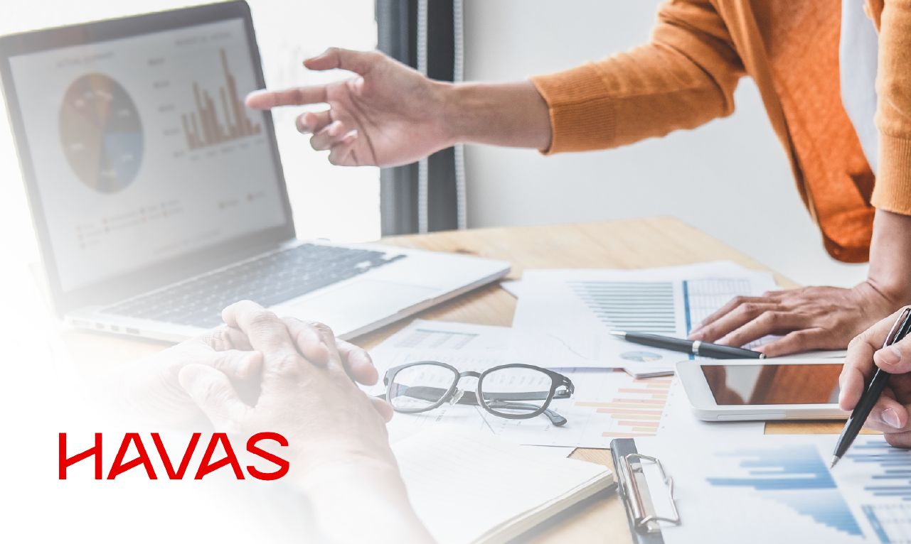 Duas pessoas analisam gráficos e relatórios numa secretária com portátil, tablet, documentos e óculos, com o logótipo da Havas em destaque.