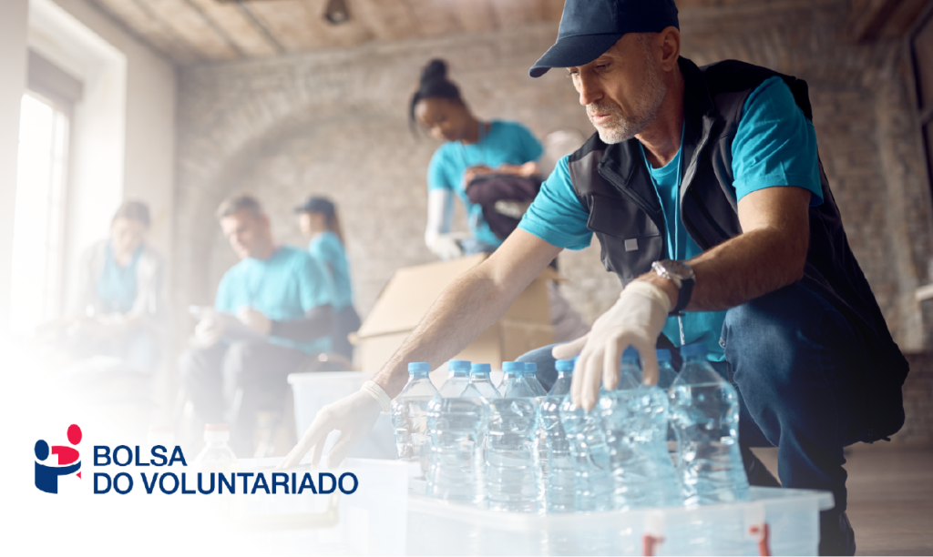 Grupo de voluntários a organizar bens essenciais, com destaque para um homem a acondicionar garrafas de água. O logótipo da Bolsa do Voluntariado reforça o espírito de solidariedade, ação social e impacto comunitário.