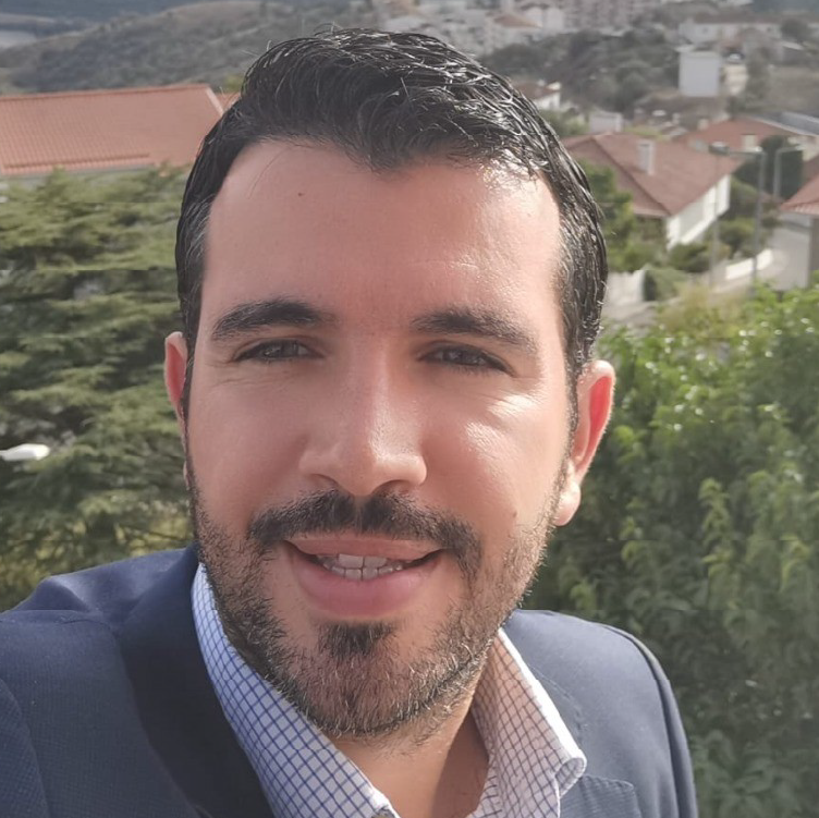 Homem com cabelo escuro e barba aparada, a sorrir, vestido com blazer azul e camisa aos quadrados, em ambiente exterior com casas e vegetação ao fundo.
