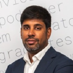 Homem com cabelo e barba curta escura, vestido com blazer azul e camisa branca, em frente a um fundo com palavras parcialmente visíveis.