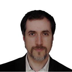Homem com cabelo escuro e barba grisalha, a olhar para a câmara com expressão neutra, vestido com blazer preto e camisa branca, sobre fundo branco liso.