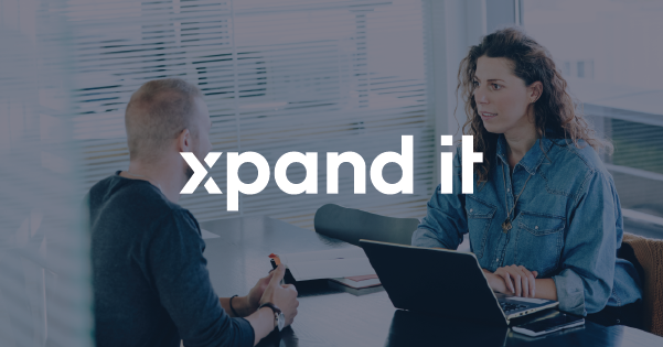 Candidate and recruiter in conversation in a modern office, with a laptop on the table. The Xpand IT logo is overlaid at the center of the image. Banner used to promote an open position for a Talent Acquisition Specialist.