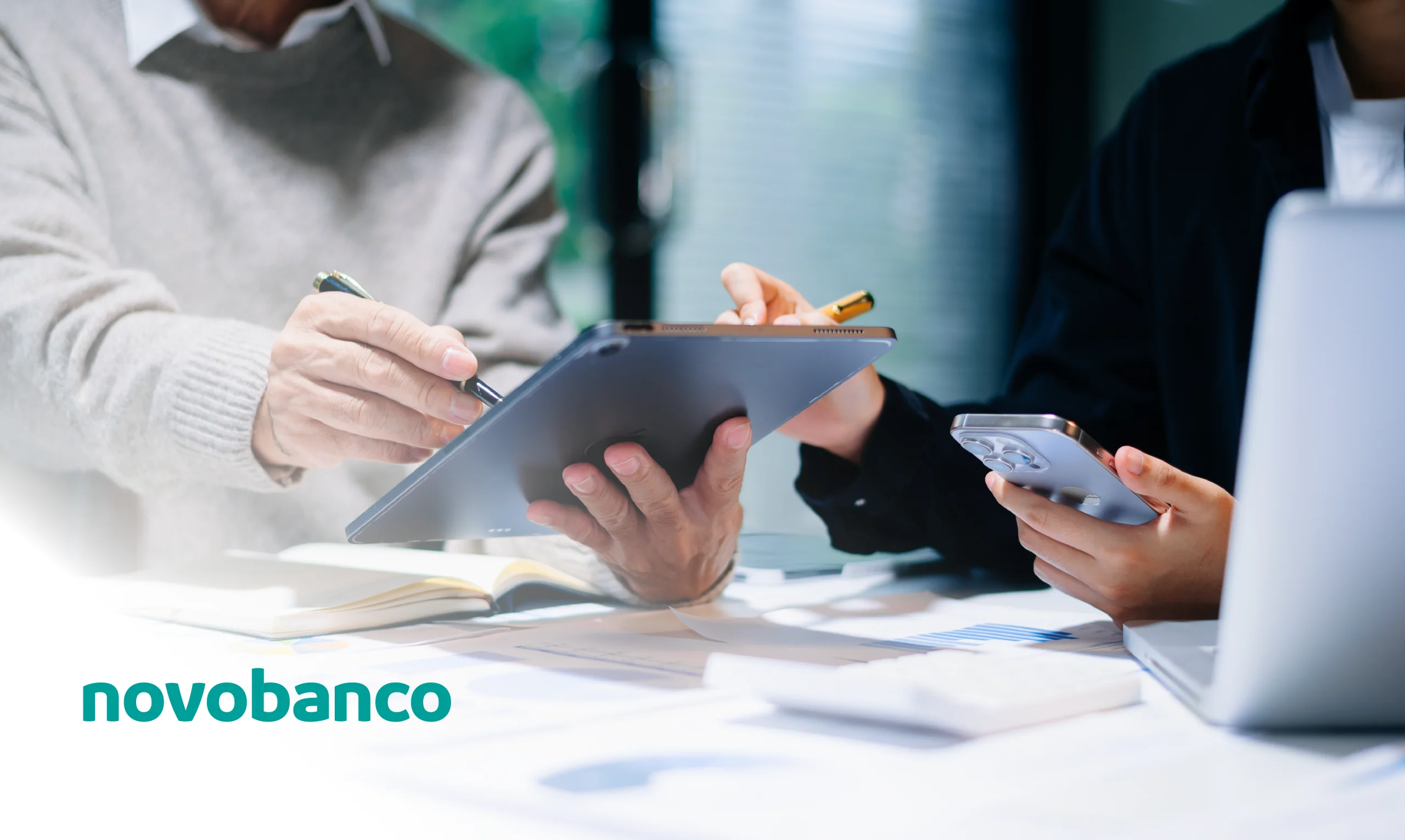 Two people collaborating in an office, using a tablet and a smartphone on a desk with documents and a laptop; novobanco logo in the lower-left corner.