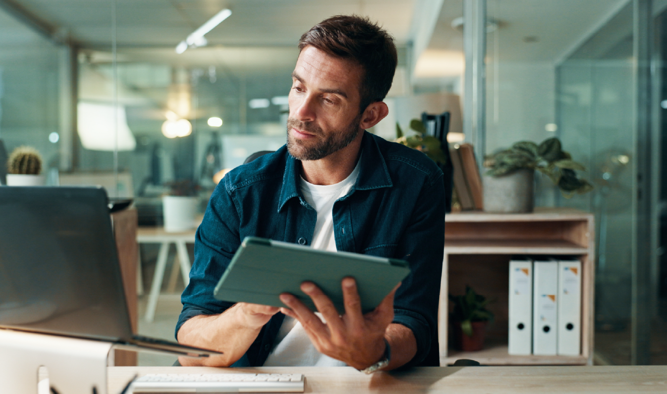 Profissional sentado num escritório moderno utiliza um tablet enquanto consulta um computador portátil, ilustrando processos de decisão, produtividade e adoção de tecnologia em ambientes de trabalho digitais.