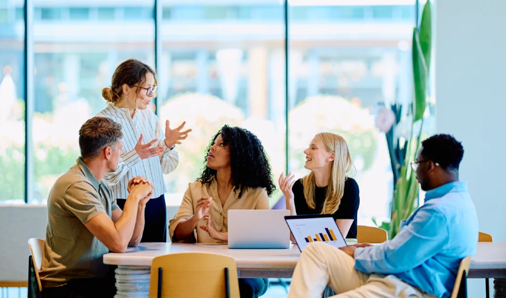 Equipa multidisciplinar reunida à volta de uma mesa, a colaborar e discutir ideias com portáteis num espaço de trabalho moderno.